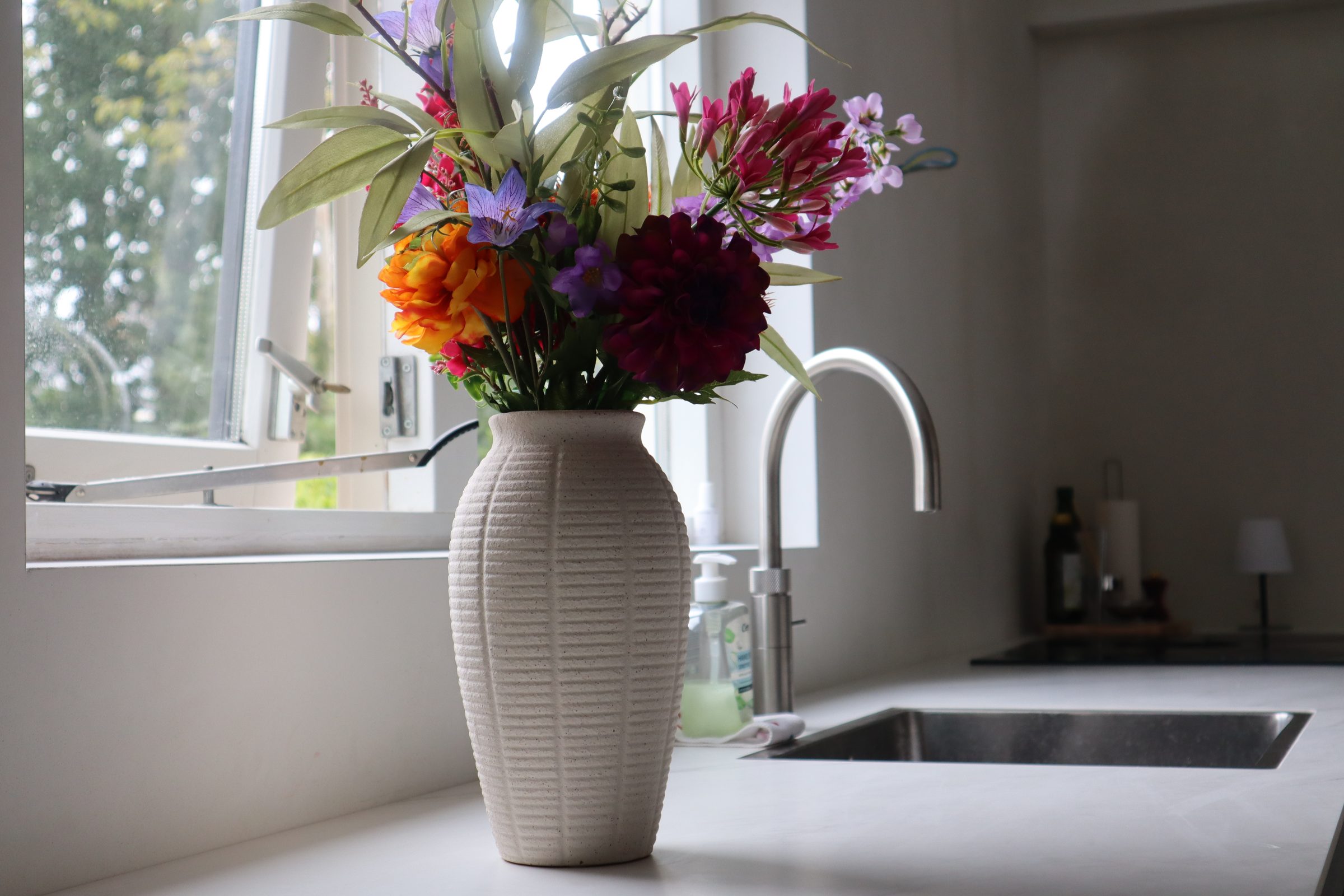 Kleurrijk boeket in een witte vaas bij het raam van een lichte keuken